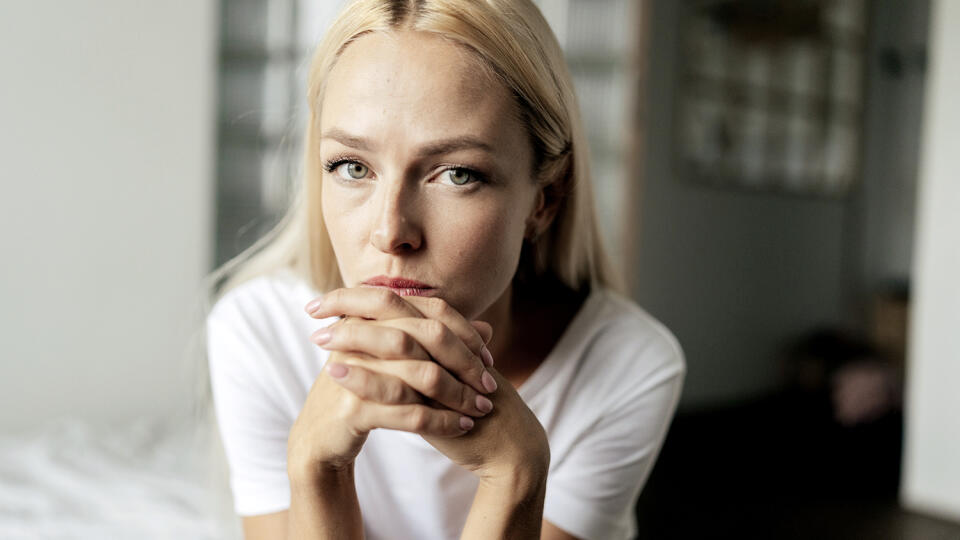 Close up face of thoughtful blonde female sitting alone in living room and serious looking at camera holding hands on chin, thinking over health problems, feeling sadness, boredom, apathy.