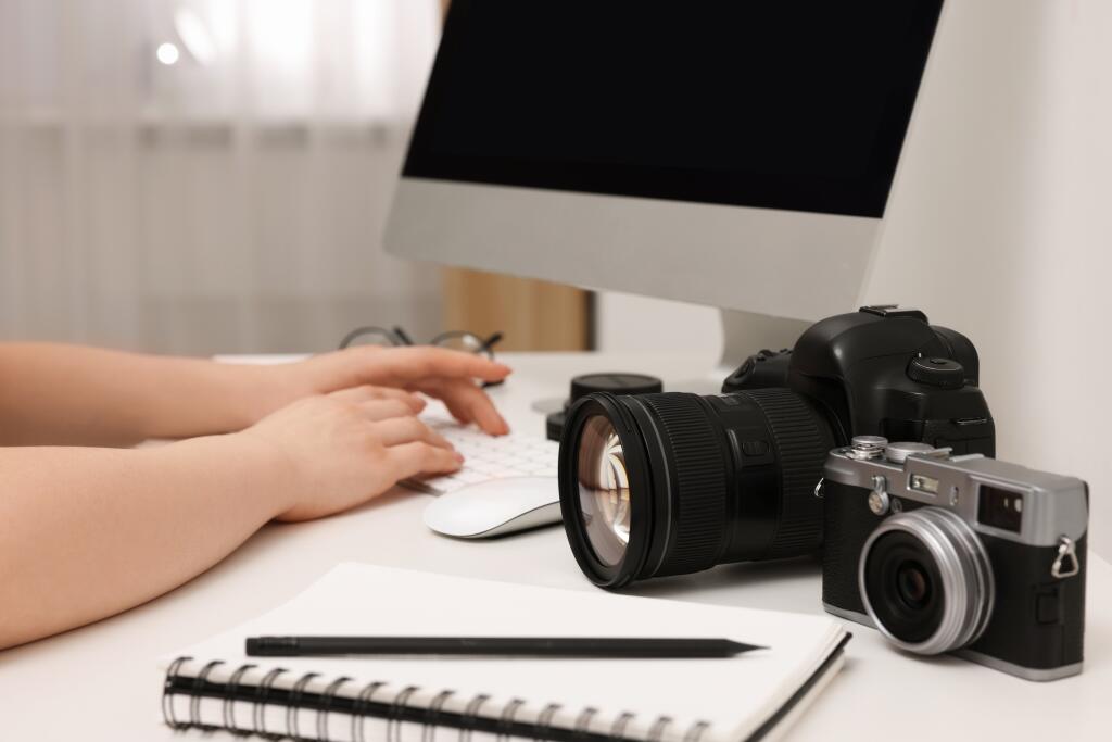 Photographer,Working,On,Computer,At,White,Table,With,Cameras,Indoors,