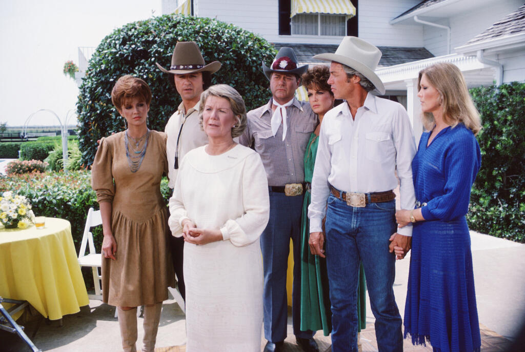 In a scene from the American television series 'Dallas, cast members gather on the porch of a large house, Dallas, Texas, 1981. From left, American actors Victoria Principal (as Pamela Barnes Ewing), Patrick Duffy (as Bobby Ewing), Barbara Bel Geddes (as Eleanor Southworth 'Miss Ellie' Ewing) (foreground), Larry Hagman (as John Ross 'J.R.' Ewing, Jr.), Linda Gray (as Sue Ellen Ewing), Steve Kanaly (as Ray Krebbs), and Susan Howard (as Donna Culver). (Photo by CBS Photo Archive/Getty Images)