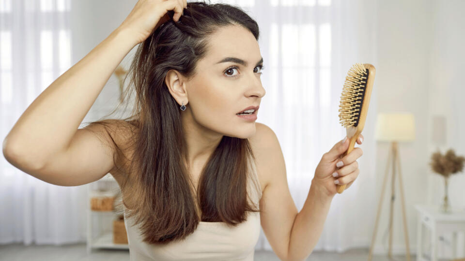 Portrait of woman in her 20s or 30s looking at her reflection with scared nervous expression as she notices bad signs like scalp dandruff, hair thinning, or hair falling out. Hair loss problem