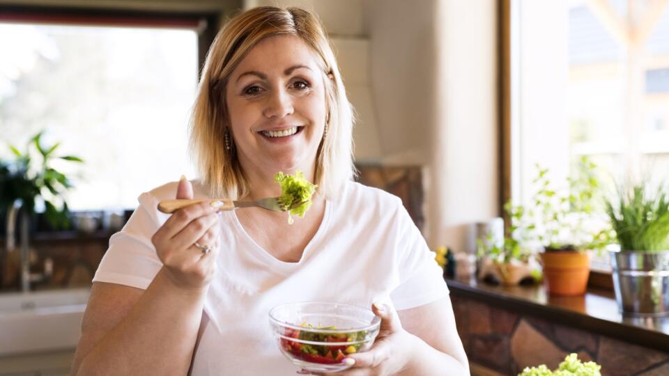 Attractive blonde overweight woman in white t-shirt at home eating a delicious healthy vegetable salad in her kitchen.