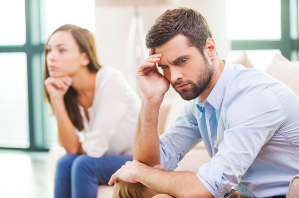 Relationship breakdown. Depressed young man holding hand on head and looking away while woman sitting behind him on the couch