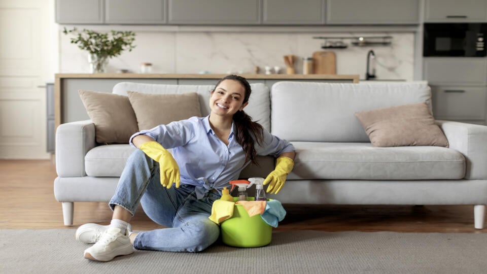 Content woman in casual attire and yellow gloves sitting on the floor, taking break with bucket of cleaning supplies next to her, living room interior, free space