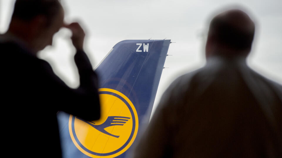 Dvaja muži sa pozerajú na lietadlo spoločnosti Lufthansa cez sklo v letiskovej hale vo Frankfurte.