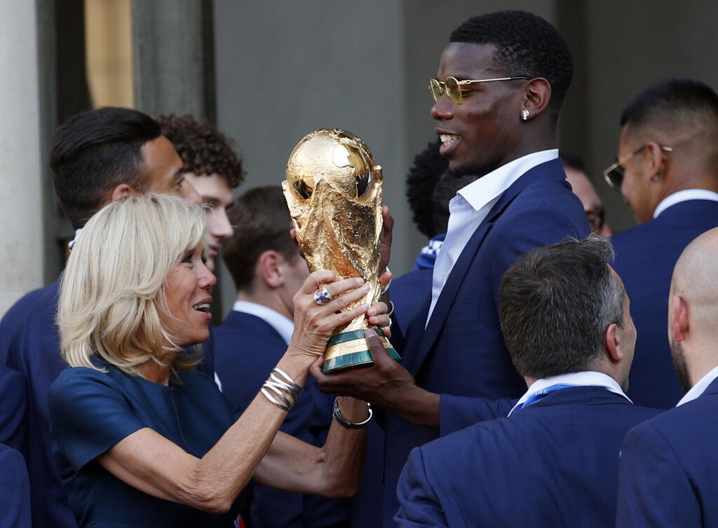 Na snímke francúzsky futbalový reprezentant Paul Pogba (vpravo) pomáha zdvihnúť trofej majstrov sveta manželke francúzskeho prezidenta Brigitte počas prijatia u francúzskeho prezidenta Emmanuela Macrona po príchode do Paríža 16. júla 2018.