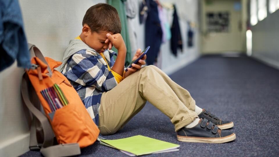 Little,Boy,With,Backpack,Holding,Smartphone,Sitting,Alone,In,School