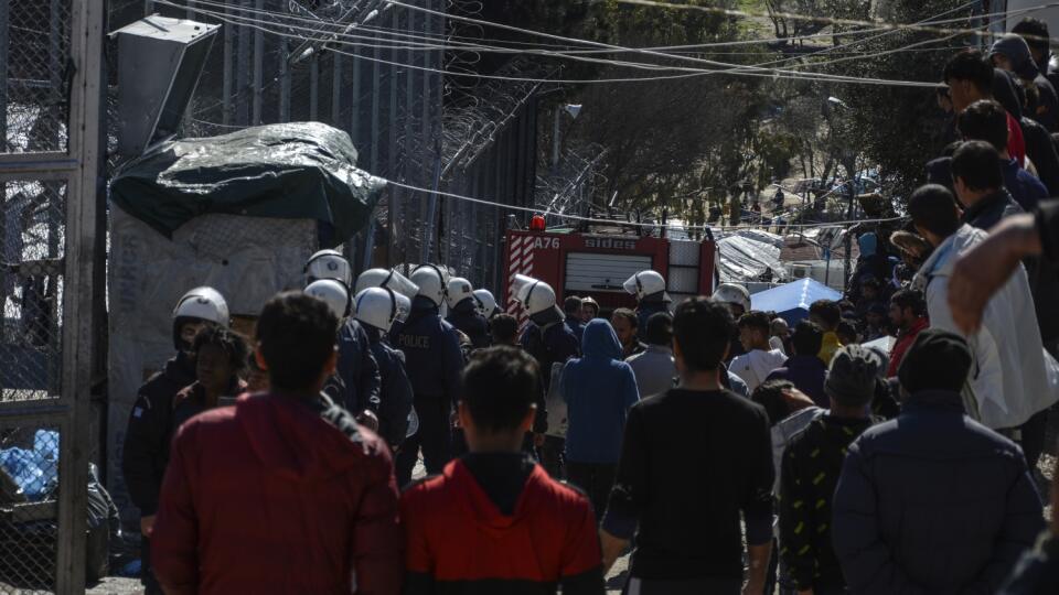 JB 19 Lesbos - Migranti sa zhromažďujú, zatiaľčo policajti hliadkujú pred bránou v utečeneckom tábore Moria na severovýchodnom gréckom ostrove Lesbos 16. marca 2020. Rozsiahly požiar vypukol v pondelok v utečeneckom tábore Moria na gréckom ostrove Lesbos. Príčina požiaru podľa hasičov nie je známa a nehlásili ani obete na životoch. Podľa svojich vyjadrení však mali ťažkosti pri hasení požiaru, pretože dočasné ubytovacie zariadenia pre migrantov sa nachádzajú v jeho tesnej blízkosti. FOTO TASR/AP