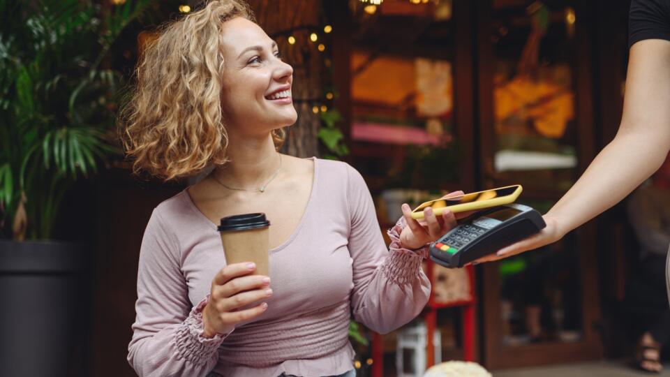 Young smiling woman 20s in casual clothes at cafe buy breakfast sit at table hold wireless bank payment terminal mobile phone to process acquire payments relax in restaurant during free time indoors.
