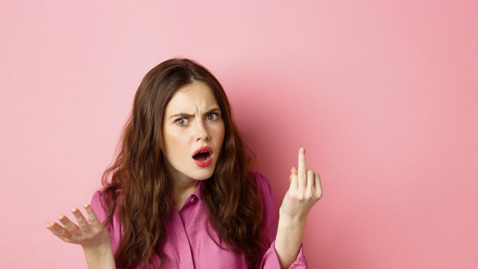 Annoyed girlfriend arguing about proposal, wants to get married, showing finger without engagement ring, standing confused against pink background.