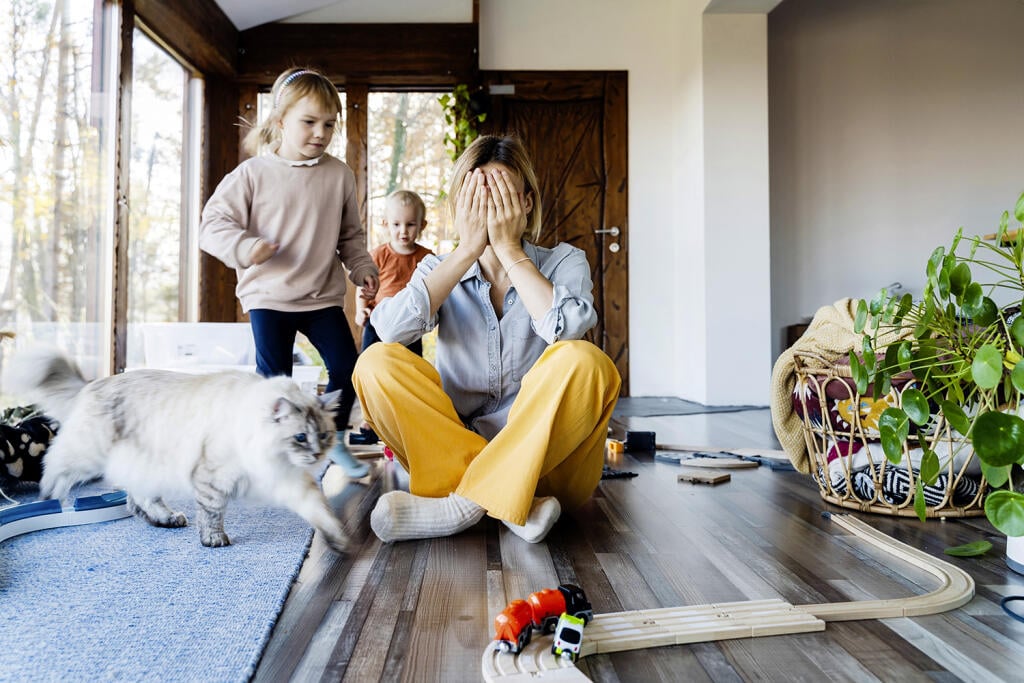 Frustrating woman alone struggling burnout with kids. Stressed out mother sitting on floor in middle of toys while children naughty running around her at room. Family home with chaos, mess.