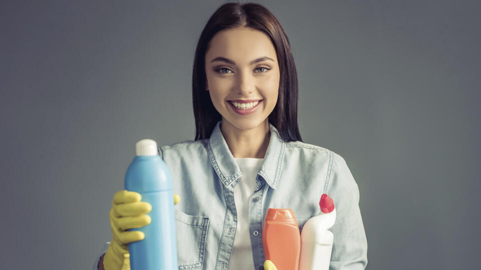 Beautiful young woman in protective gloves is holding detergents, looking at camera and smiling, on gray background