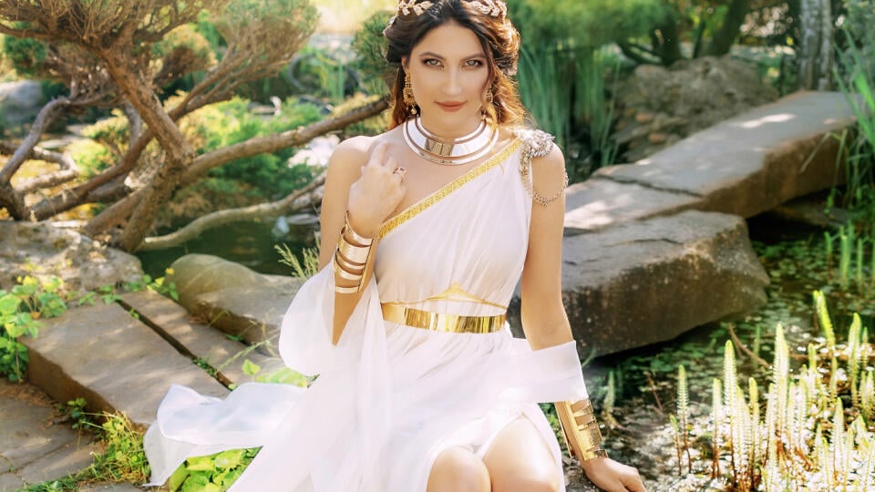 Art photo Fantasy woman greek goddess sits in spring garden green tree grass nature day magic sun light. Sexy girl queen antique style white silk dress, gold jewelry set necklace bracelet crown belt