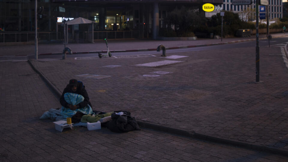 JB 15 Tel Aviv - Bezdomovec sedí na ostrovčeku na križovatke pri bilborde vyzývajúcom ľudí, aby nosili ochranné rúška počas národného lockdownu v Tel Avive 8. januára 2021. V Izraeli je v platnosti v poradí už tretí lockdown, ktorý má spomaliť opätovné šírenie koronavírusu. Na dodržiavanie obmedzení pohybu obyvateľstva dohliada v celej krajine zhruba 6000 policajtov, informovala agentúra DPA. Policajti na pouličných kontrolných stanovištiach monitorujú dodržiavanie opatrenia, v súlade s ktorým s