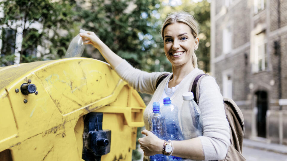 Woman throwing plastic waste, bottles into recycling container in front her apartment. Woman sorting the waste according to material into colored bins.