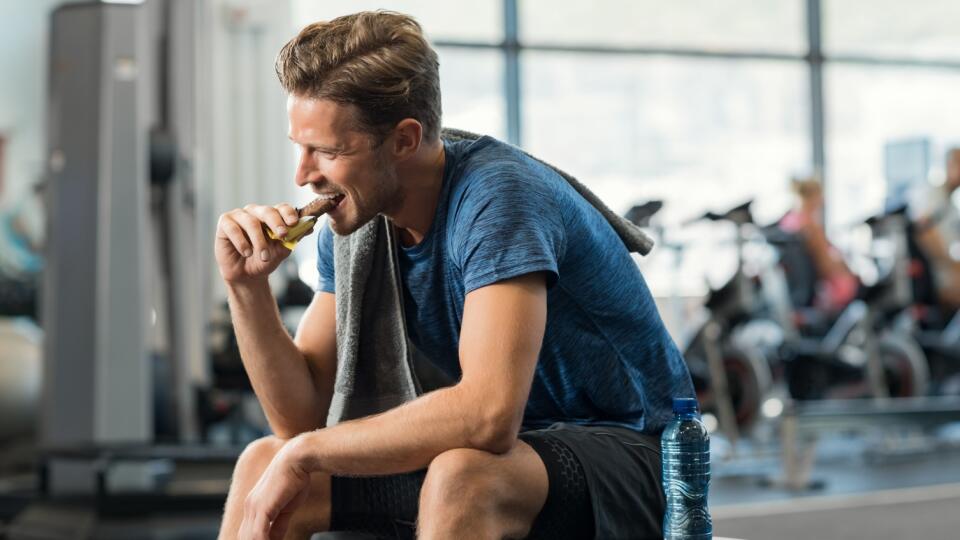 Sweaty young man eating energy bar at gym. Handsome mid guy enjoying chocolate after a heavy workout in fitness studio. Fit man biting a snack and resting on bench.