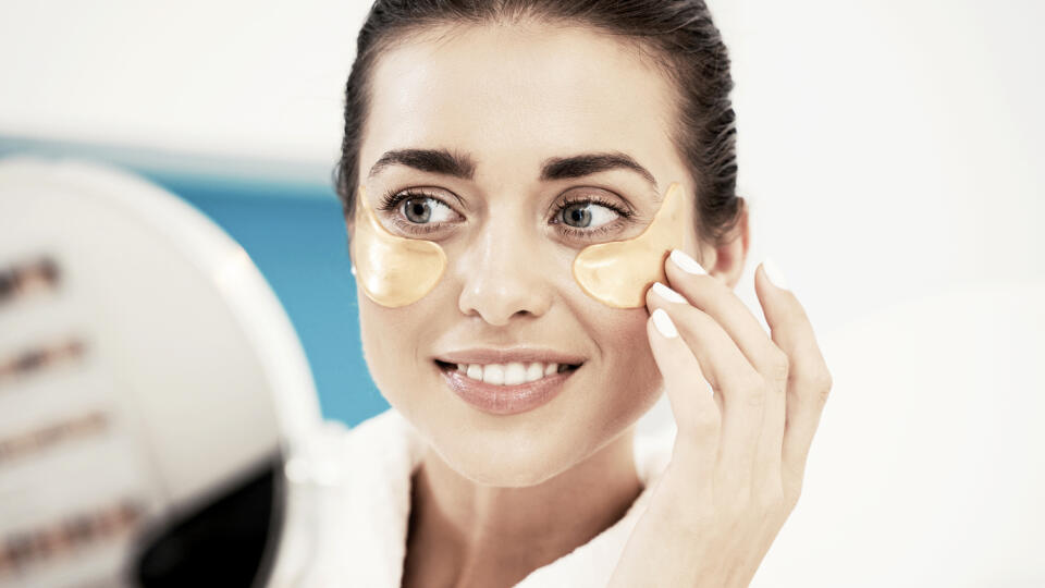 Smiling young brunette woman wearing bathrobe applying cosmetic eye patches while standing at the bathroom mirror