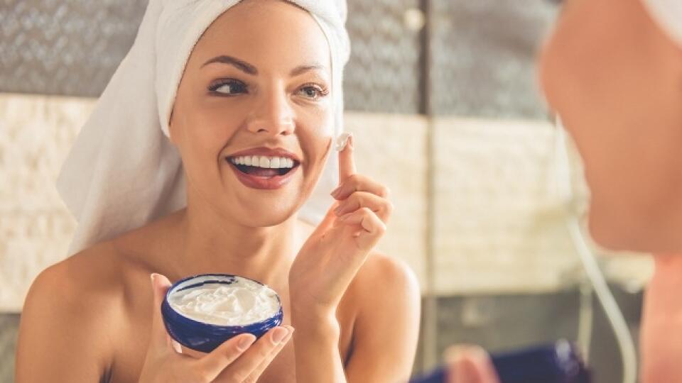 Beautiful young woman in bath towel is applying cream on her face and smiling while looking into the mirror in bathroom