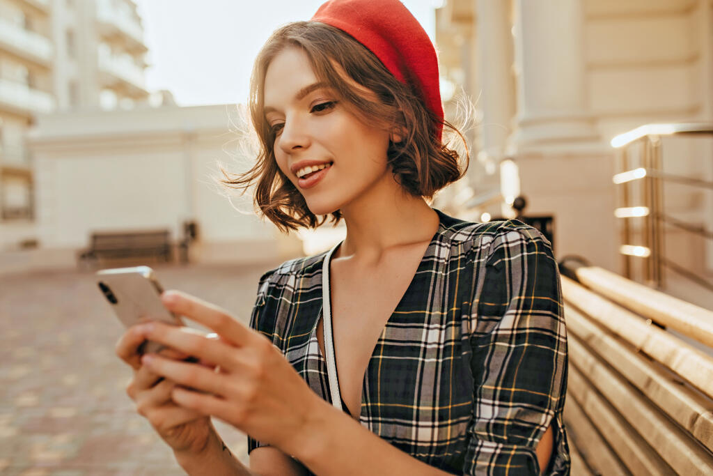 Winsome elegant girl texting message in autumn day. Debonair short-haired woman in red beret standing on the street with phone.