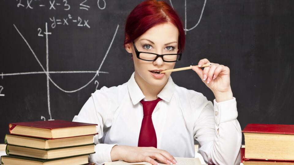 Beautiful redhead teacher sitting behind a desk in a classroom. She is wearing a white shirt, bright red necktie and a pair of glasses. Mathematical symbols on the blackboard behind her. Several books on the desk in front of her. 