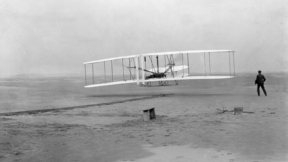 Kill Devil Hill, 17. decembra 1903. Historický okamih – prvé motorové lietadlo sa vznáša. Orville Wright ho pilotuje, Wilbur beží vedľa a kontroluje priebeh pokusu.