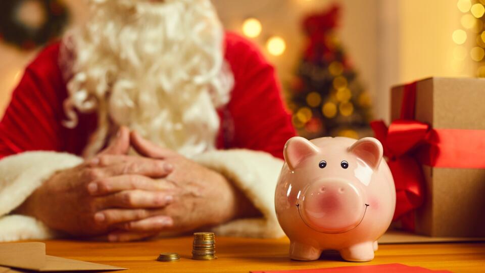 Close-up of piggy bank on desk with presents and Christmas charity event invitations with old Santa Claus in blurred background. Donation to orphan funds, saving up money, opening bank account concept