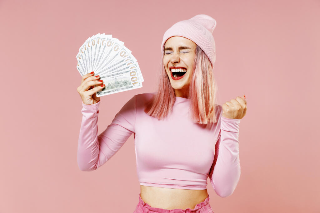Young woman with dyed rose hair in rosy top shirt hat holding fan of cash money in dollar banknotes do winner gesture isolated on plain light pastel pink background. People lifestyle fashion concept