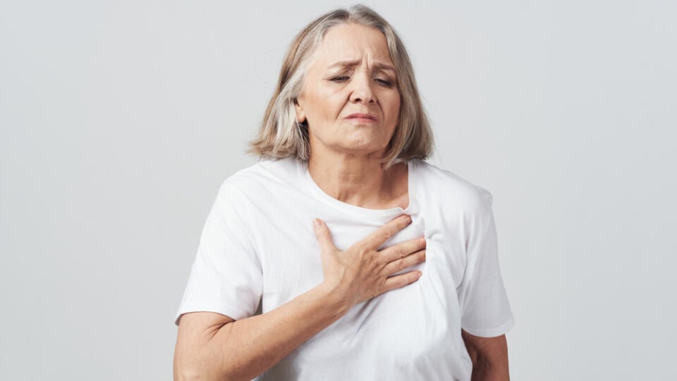 Ill,Elderly,Woman,Holding,Her,Chest,Against,A,Gray,Background