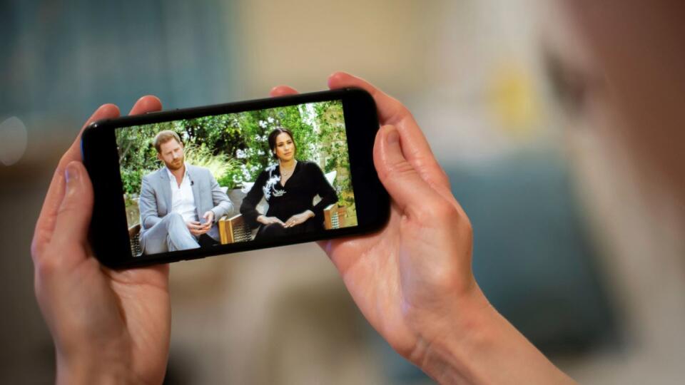 A man watches a phone screen showing an interview of Prince Harry and Meghan, The Duchess of Sussex, by Oprah Winfrey